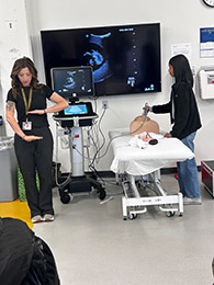 HEROES students get ultrasound demonstration on a simulation mannequin, featuring instructors using diagnostic imaging equipment in a clinical skills lab.